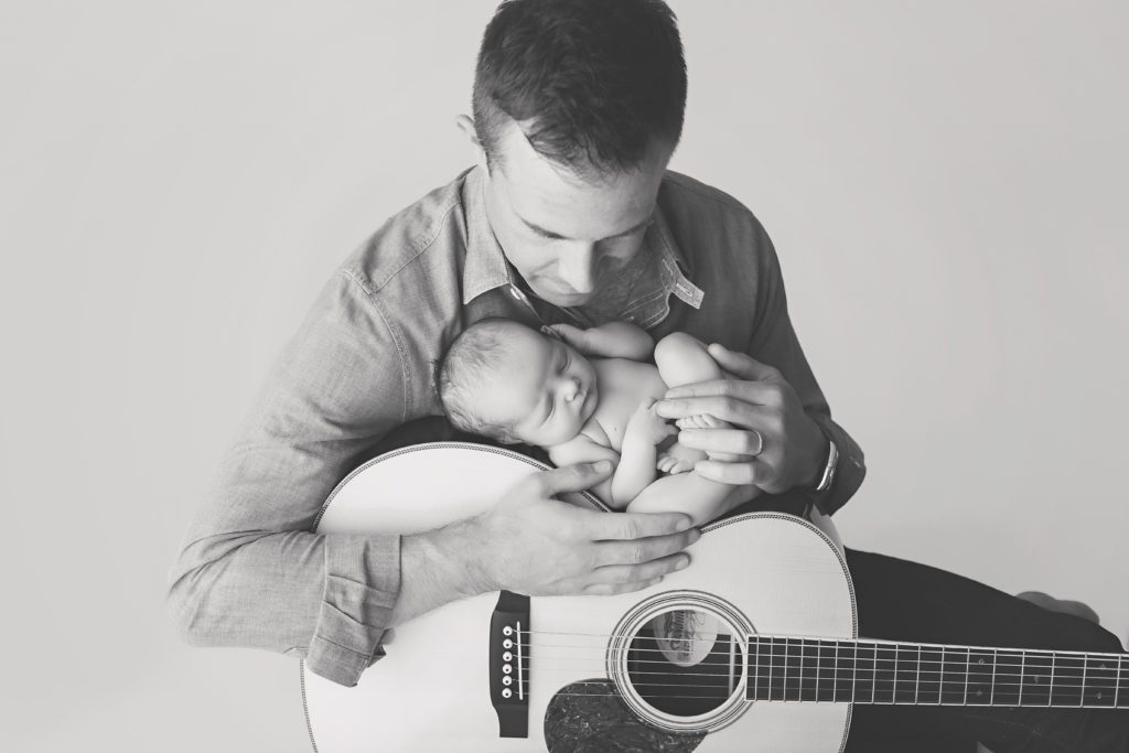black and white image of dad holding newborn baby and guitar