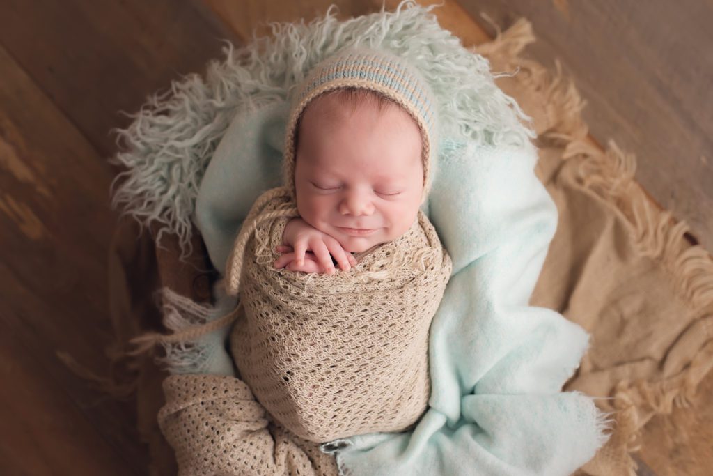 newborn boy wrapped with a bonnet and smiling light blue and tan colors