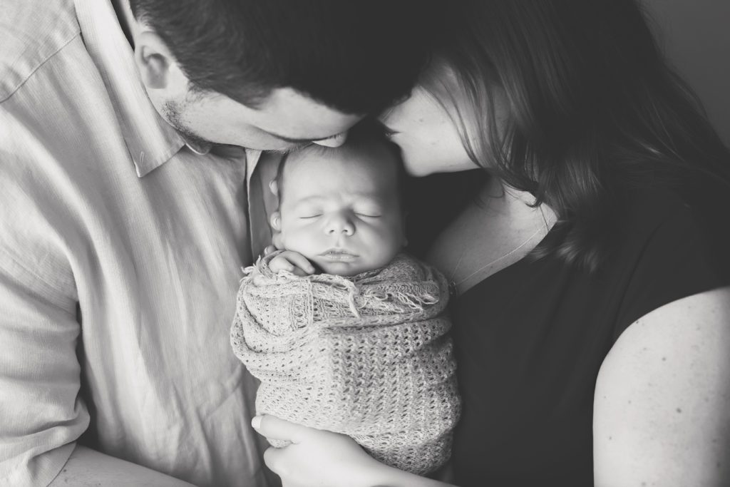 black and white image of new mom and dad holding newborn baby boy and kissing him on the head