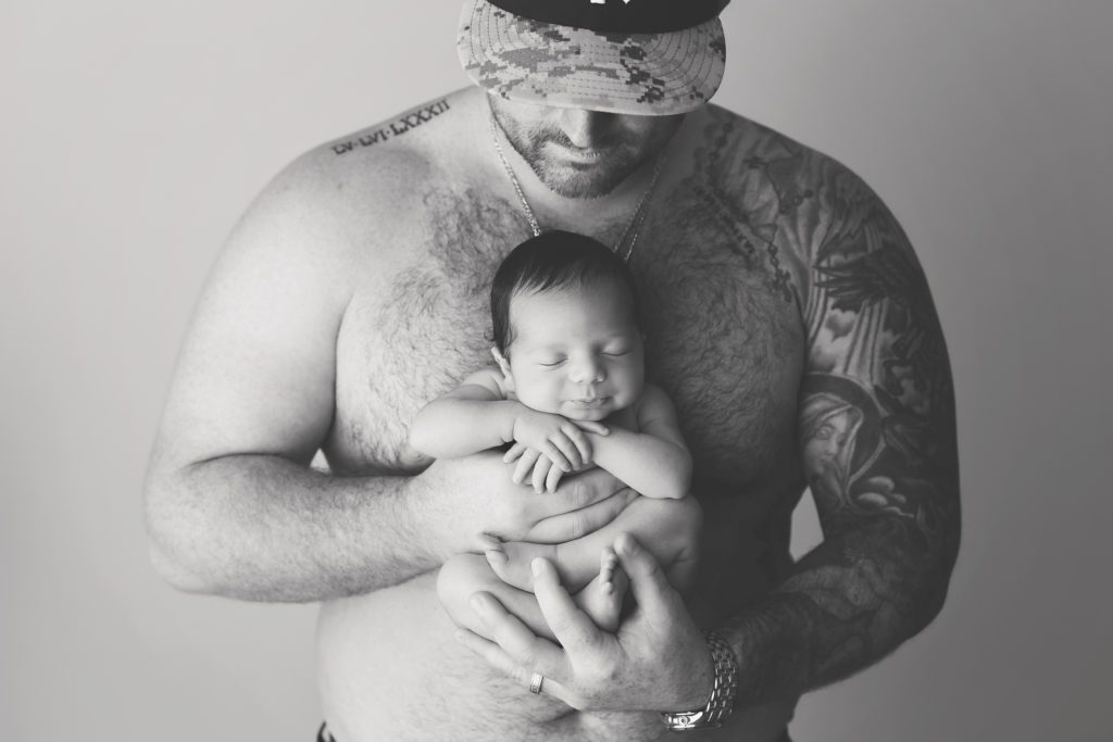 portrait of new dad with no shirt and tattoos holding baby boy 
