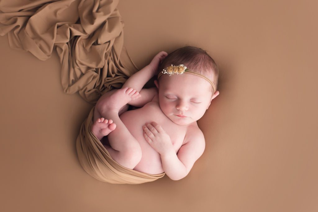 newborn baby girl wrapped in a carmel color wrap on background and posed simply with matching tieback