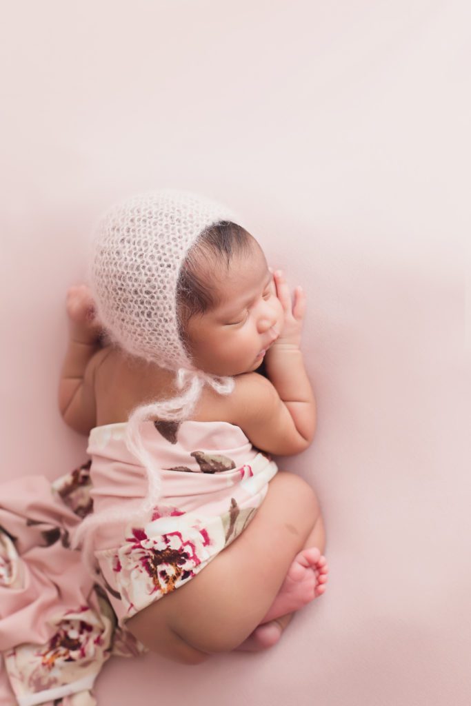 newborn baby girl posed on pink with pink bonnet and floral wrap