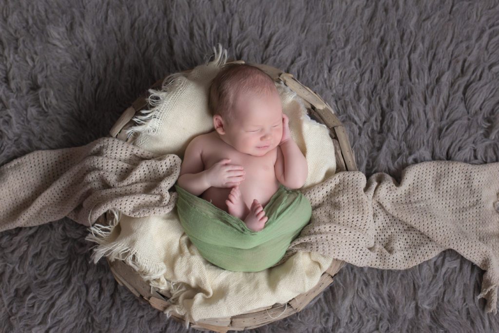newborn baby boy laying in round bowl on gray rug smiling
