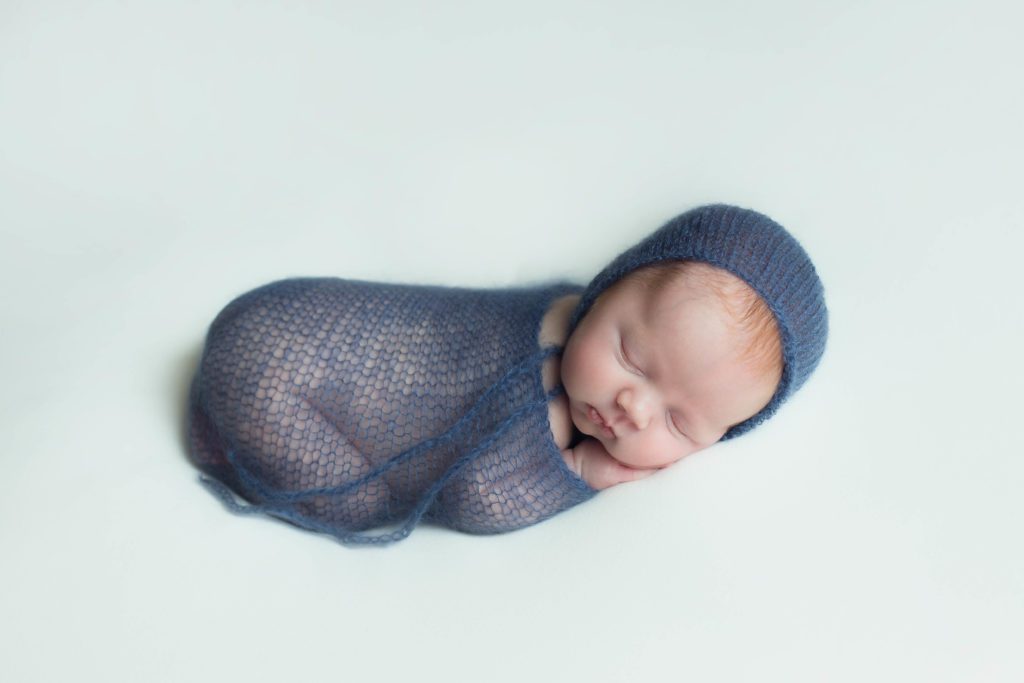 red headed baby boy wrapped in blue wrap and bonnet laying on white background 