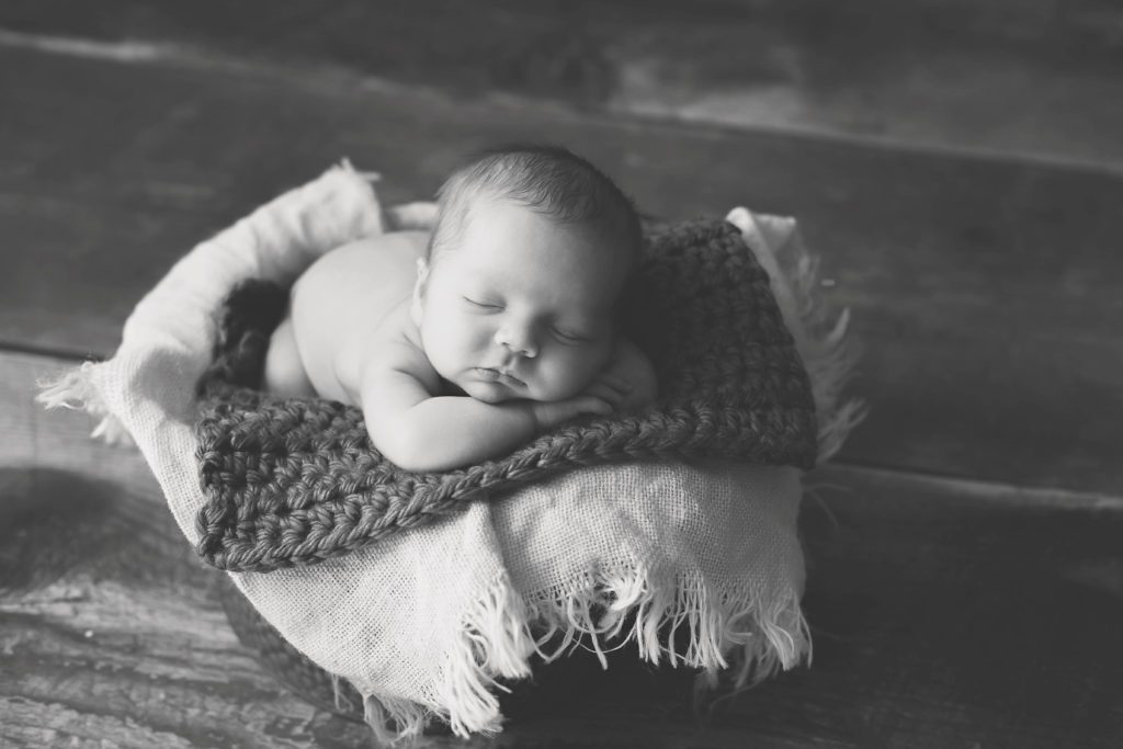black and white photo of baby laying in a basket with blankets on wood floor