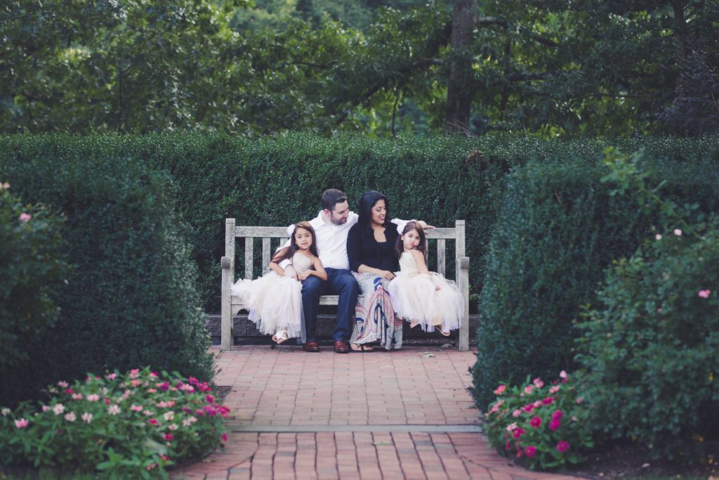 wide shot of family sitting on park bench among beautiful green trees and shrubs and flowers Alyssa Joy Photography