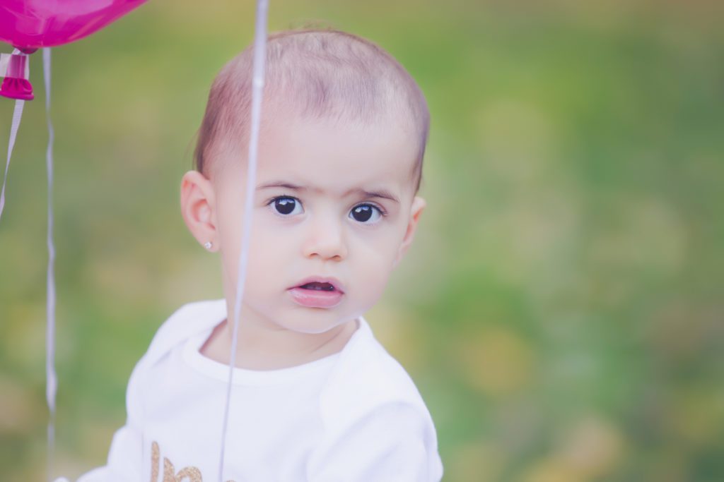 one year old girl looking at camera from behind pink balloons