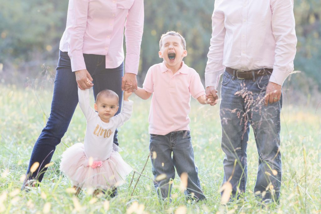family of four posing in a grassy field with focus on one year old little girl and older son dressed in pink