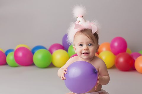 photo of one year old girl holding purple balloon with pink birthday hat and balloons