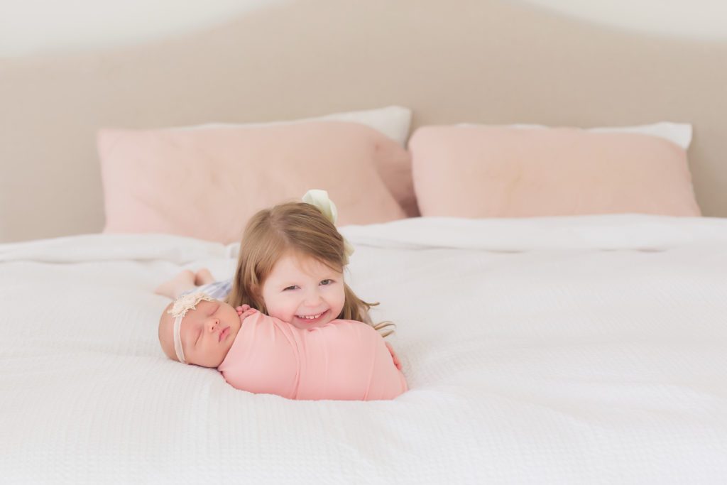 lifestyle newborn session with baby girl and her 3 year old big sister on bed with white and pink