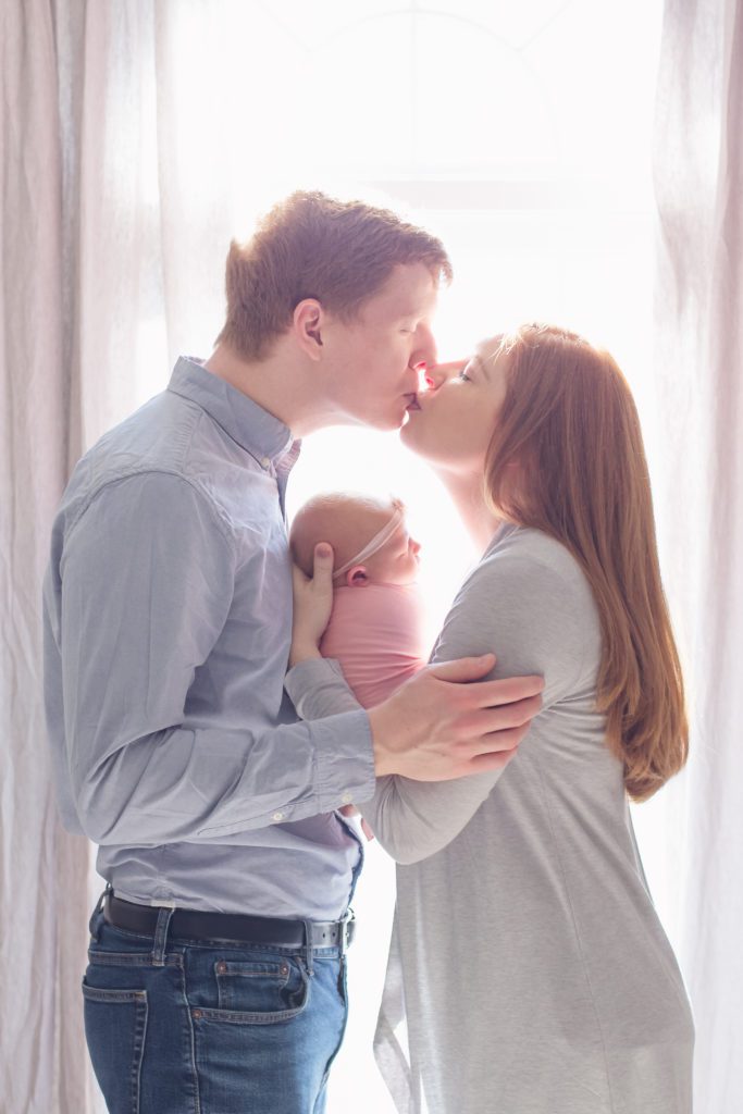 picture of mom and dad kissing in front of window while holding newborn baby girl Alyssa Joy Photography