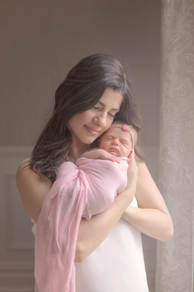 new mom holding newborn baby girl wrapped in pink by window in her home at lifestyle newborn photo session