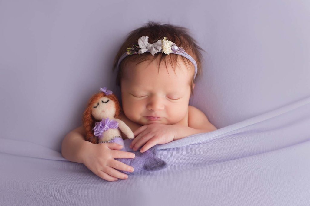red headed baby girl laying down facing up holding mermaid doll