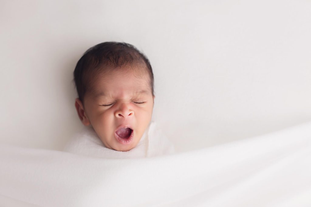 tucked in newborn boy yawning wrapped in white during baby photography session with Alyssa Joy